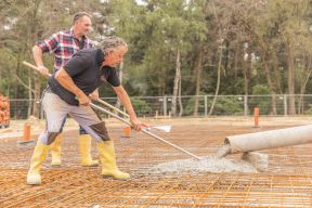 Betonarbeiten Zwei Mitarbeiter verstreichen Beton, das aus einem Rohr kommt, für ein neues Fundament.