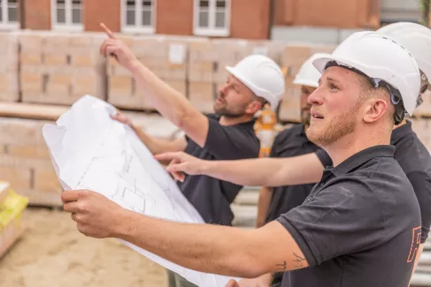 Vier Maurer halten einen Bauplan und schauen in die Ferne.