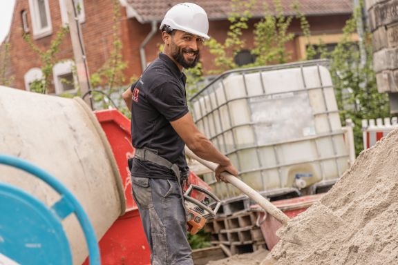 Ein Bauhelfer l&auml;chelt in die Kamera w&auml;hrend er mit einer Schaufel Zement in einen Mischer kippt.