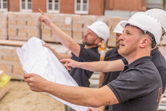Vier Maurer halten einen Bauplan und schauen in die Ferne.