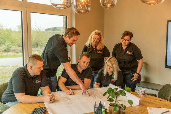 Impression_11 Nico Robben erklärt den Mitarbeitern etwas auf einem Bauplan, der auf einem Tisch liegt.