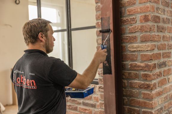 Ein Mitarbeiter streicht einen Eisenpfosten zur Sanierung mit schwarzer Farbe an.